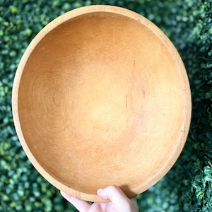 May include: A light brown, wooden bowl with a rounded shape. The bowl has a smooth, natural finish and appears to be hand-carved. The bowl is held by a hand, showcasing its size and form. The bowl is likely used for serving or display.