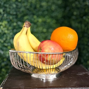 May include: A silver wire fruit bowl filled with fresh fruit. The bowl contains yellow bananas, a red apple, and a bright orange. The bowl is sitting on a dark surface with a green background.