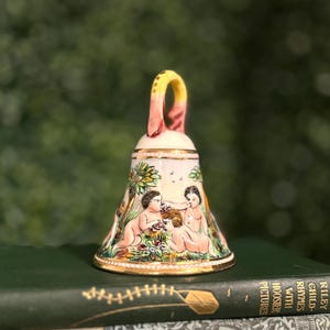 May include: A decorative bell with a handle, featuring a painted scene of cherubic figures in a garden setting. The bell has a gold rim and a cream-coloured base. The handle is yellow and pink. The bell is sitting on top of a stack of books.