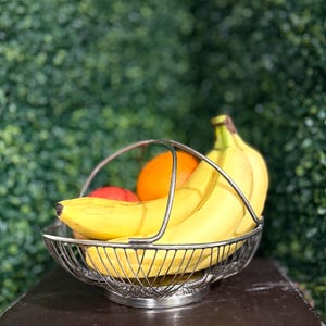 May include: A silver wire fruit basket filled with ripe bananas, an orange, and a red apple. The basket has a handle and sits on a dark surface, with a green hedge in the background.