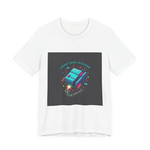 Puede incluir: Camiseta blanca con un diseño de consola de videojuegos retro con propulsores de cohete, corazones y estrellas. El texto "Latest Level Unlocked!" está impreso en turquesa sobre la consola. El diseño está sobre un cuadrado gris oscuro.