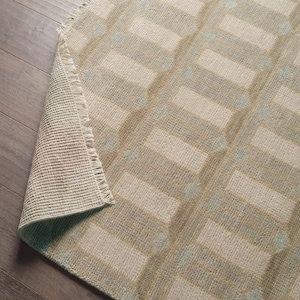 May include: A rectangular area rug with a geometric pattern in shades of beige, light blue, and tan. The rug's design features repeating rectangular shapes and small circular accents. The edge of the rug is frayed.