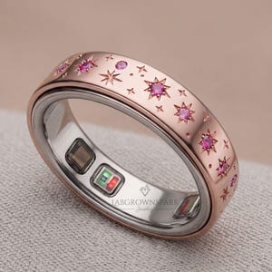 May include: A rose gold smart ring featuring a celestial pattern of engraved starbursts and inlaid pink gemstones. The interior band is silver-toned with visible electronic sensors and the text LABGROWNSPARK Jewellery printed in black.