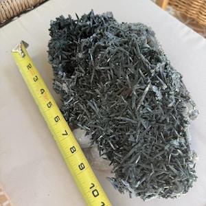 Puede incluir: Un racimo mineral de color gris oscuro con forma de aguja que presenta numerosos cristales afilados y alargados. Una cinta métrica amarilla está colocada junto al espécimen, indicando una longitud de unos 30,5 cm para la formación.