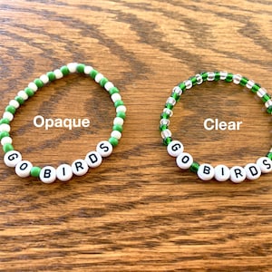 May include: Two beaded bracelets with the words "GO BIRDS" spelled out in black letters on white beads. The bracelet on the left has opaque green and white beads, while the bracelet on the right has clear green and white beads.