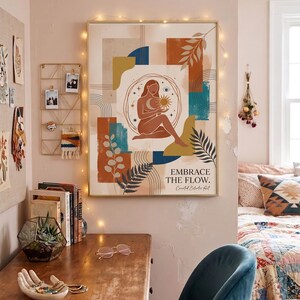 May include: A framed art print with a gold frame featuring an abstract design with a woman holding a sun and moon. The artwork includes the text "EMBRACE THE FLOW." The color palette includes shades of brown, blue, and orange. The print hangs above a wooden desk.