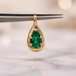 Puede incluir: Un colgante en forma de lágrima con una piedra preciosa de color verde vibrante engastada en un marco de metal dorado pulido. El colgante está sujeto por unas pinzas metálicas, mostrando el corte facetado de la piedra y sus garras.