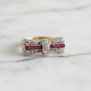 May include: A gold and silver ring featuring a central row of square-cut red rubies flanked by two rows of round brilliant-cut diamonds. The design includes a vertical band of three diamonds in the center, creating a bow-like shape across the finger.