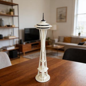 May include: A cream-colored model of the Seattle Space Needle, featuring a distinctive saucer-shaped top with black accents and a tall, open-frame tower. The model is displayed on a dark wooden table, showcasing its architectural details.