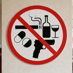 Könnte beinhalten: Rundes weißes Schild mit rotem Rand und roter Diagonallinie. Das Schild zeigt schwarze Silhouetten einer Zigarette, einer Flasche, eines Glases, Pillen und einer Pistole, alle durchgestrichen.