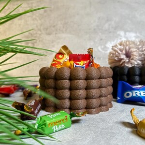 Könnte beinhalten: Eine braune, strukturierte, kuchenartige Struktur, gefüllt mit verschiedenen Süßigkeiten, darunter ein Schokoriegel und verpackte Pralinen. Eine blaue Oreo-Packung und grüne verpackte Süßigkeiten sind ebenfalls zu sehen.