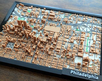 May include: A 3D-printed map of Philadelphia, showing detailed building and street layouts. The map is framed in black, with the city name in white. Buildings are light brown, and parks are green. The map is in a black frame.