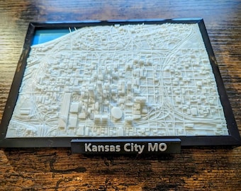 May include: A 3D-printed white map of Kansas City, MO, framed in black. The map shows the city's layout with detailed streets and buildings. A small black sign at the bottom reads "Kansas City MO".