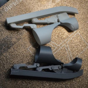 May include: Two grey 3D-printed objects, possibly components of a mechanism, are shown. The top object is light grey and has a hinged design. The bottom object is dark grey and has a curved base and a similar hinged structure.