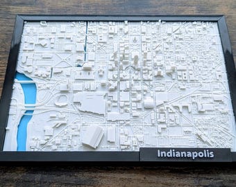 May include: A 3D-printed white map of Indianapolis, Indiana, in a black frame. The map shows detailed buildings, streets, and a blue river. The word "Indianapolis" is printed on a black base, a city in the United States.