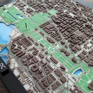 Peut inclure: Une carte imprimée en 3D de Washington, DC, encadrée en noir. La carte présente des modèles de bâtiments bruns détaillés, des espaces verts et un plan d'eau bleu. Le texte "Washington DC" est imprimé sur le cadre.