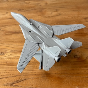 May include: A gray model airplane, a fighter jet, is displayed on a wooden surface. The jet has detailed wings, tail fins, and engine nozzles. The model is a detailed replica of a military aircraft.