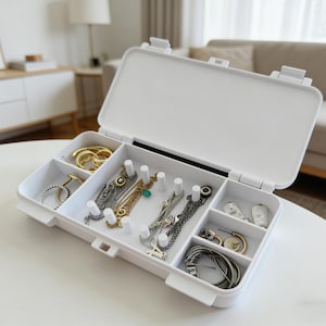 May include: A white, open jewelry organizer with multiple compartments. The organizer contains various pieces of jewelry, including gold and silver necklaces, rings, and earrings. The organizer is on a white surface, and the background includes a couch and a lamp.