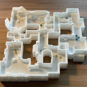 Puede incluir: Un modelo blanco impreso en 3D de un diseño de edificio, con múltiples habitaciones y pasillos. Pequeños objetos dorados y azules se colocan dentro de la estructura. El modelo está sobre una superficie de madera.