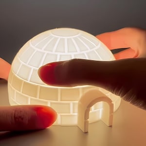 Puede incluir: Un iglú de cerámica blanca brillante con una parte superior redondeada y una entrada arqueada. El iglú está iluminado desde dentro, y la imagen muestra el iglú sostenido por dedos.