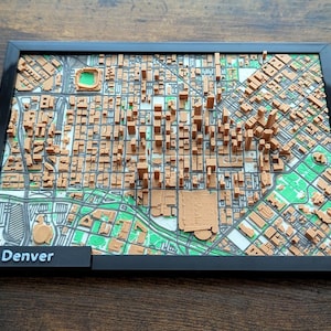 May include: A framed 3D map of Denver, Colorado, featuring a detailed representation of the city's layout. The map is in shades of brown and green, with a black frame and the city name displayed on a small black plaque.
