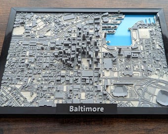 May include: A 3D-printed map of Baltimore, Maryland, in a black frame. The map is gray with a blue section representing the harbor. The city name "Baltimore" is displayed on a black base at the bottom of the frame. The map shows detailed city features.
