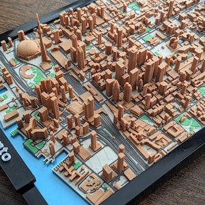 Puede incluir: Un mapa impreso en 3D de Toronto, Canadá, con edificios y monumentos en miniatura detallados en tonos marrones y beige. El mapa está sobre un fondo azul y blanco, con el nombre de la ciudad en letras blancas sobre un marco negro.
