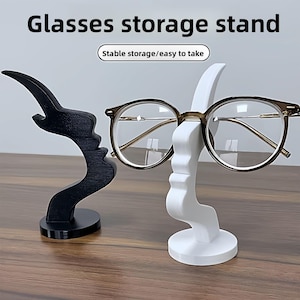 May include: A white and a black glasses storage stand, each shaped like a stylized face. The white stand holds a pair of eyeglasses with brown frames. Text on the image reads "Glasses storage stand" and "Stable storage/easy to take."