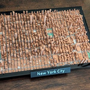 Puede incluir: Un modelo impreso en 3D de la ciudad de Nueva York, con edificios en miniatura detallados en tonos marrones. El modelo está enmarcado en negro, con un pequeño letrero que dice "New York City". Incluye áreas verdes que representan parques.