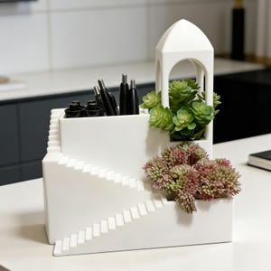 May include: A white ceramic desk organizer with a staircase design. It features compartments for pens and small items, a small arched structure, and decorative faux succulents in shades of green and pink.