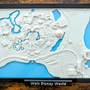 May include: A framed, three-dimensional map of Walt Disney World, featuring white raised relief details against a light blue background. The map is encased in a black frame, with the words "Walt Disney World" displayed on a small black base.