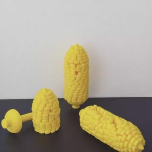 Puede incluir: Tres objetos amarillos con forma de maíz. Uno es una mazorca de maíz entera, otro es media mazorca y el tercero es media mazorca con un mango. Los objetos están sobre una superficie oscura con un fondo blanco.