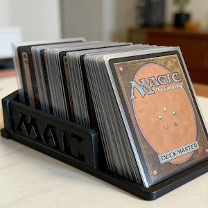 Peut inclure: Un présentoir noir avec le mot "MAGIC" découpé sur le devant, contenant une pile de cartes à collectionner Magic: The Gathering. Les cartes sont dans des pochettes de protection, avec le logo "Deckmaster" visible.