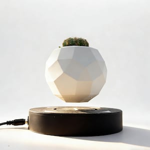 Könnte beinhalten: Ein weißer, geometrisch geformter Blumentopf schwebt über einer schwarzen Basis. Ein kleiner grüner Kaktus befindet sich in dem Pflanzgefäß. Die Basis hat ein Netzkabel. Der Hintergrund ist weiß.