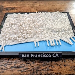 May include: A 3D model of San Francisco, CA, in a black frame. The model is white, depicting buildings and streets, with a blue section representing the bay. The text "San Francisco CA" is displayed on a black label.