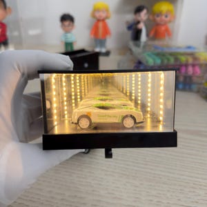 Pode incluir: Um carro de brinquedo branco com detalhes verdes e o texto "Guard the city" dentro de uma caixa de exibição retangular preta com interior espelhado e luzes LED quentes. A caixa é segurada por uma mão enluvada.