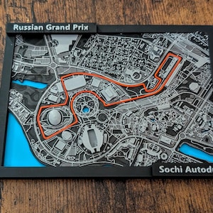 Może przedstawiać: Oprawiona mapa toru Sochi Autodrom, toru wyścigowego Grand Prix Rosji Formuły 1. Tor jest podświetlony na pomarańczowo na szarej i czarnej mapie z niebieskimi akcentami. Rama jest czarna, z napisami "Russian Grand Prix" i "Sochi Autodrom".