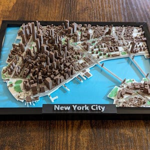 May include: A framed 3D relief map of New York City, featuring brown buildings, green parks, and blue water. The map is set against a light blue background within a black frame. A black nameplate at the bottom reads "New York City".
