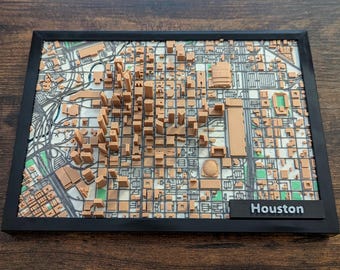May include: A framed 3D map of Houston, Texas, featuring detailed representations of buildings and streets. The map is in shades of brown, white, and green, with a black frame and a label that reads "Houston".