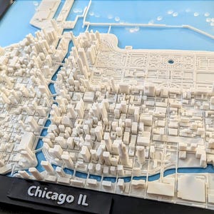 Op de afbeelding: Een 3D-reliëfkaart van Chicago, Illinois, met witte gebouwen en structuren tegen een lichtblauwe achtergrond die Lake Michigan voorstelt. De naam van de stad staat in witte tekst op een zwarte basis.