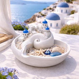 Op de afbeelding: Een witte decoratieve keramische kom met een schelpontwerp, gevuld met kleine kiezels, een zeester en een blauwe boze oog-bedel. Miniatuur witte gebouwen met blauwe koepels zijn genesteld in de schelp.