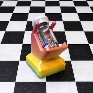 Könnte beinhalten: Ein mehrfarbiger 3D-gedruckter Halter für ein kleines Glasfläschchen. Der Halter hat einen Farbverlauf von Gelb, Orange und Rosa mit einem weißen Deckel. Das Fläschchen enthält zwei kleinere Glasröhrchen. Der Hintergrund ist ein schwarz-weißes Schachbrettmuster.