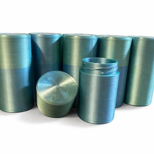 Op de afbeelding: Een verzameling cilindrische containers in blauw- en groentinten. De objecten hebben een metallic glans en een gestructureerd oppervlak. Eén container is open en onthult de binnenkant. De objecten zijn op een witte achtergrond geplaatst.