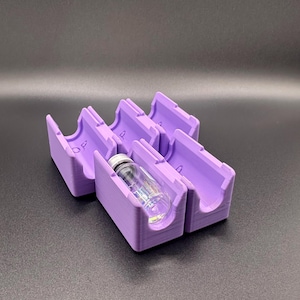 May include: A set of five lavender-colored, 3D-printed holders, each designed to cradle a small glass vial. The holders have a curved design and are arranged in a row. One vial is visible, with a silver cap.