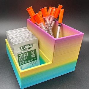 Peut inclure: Un organisateur imprimé en 3D multicolore avec des compartiments pour fournitures médicales. L'organisateur contient des seringues avec des capuchons orange et des paquets de tampons imbibés d'alcool. Les tampons sont étiquetés "CURAD" et "ALCOHOL PREP PAD".