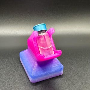 Può includere: Una piccola bottiglia di vetro con tappo blu è tenuta da un supporto rosa e viola. Il supporto ha una base rettangolare con una sfumatura da blu a viola e il supporto è di colore rosa acceso.