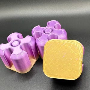 Op de afbeelding: Drie 3D-geprinte puzzelstukjes in paarse en gele tinten. De puzzelstukjes hebben een uniek, in elkaar grijpend ontwerp met een cirkelvormig midden. Het vierkante stuk is geel met een getextureerd oppervlak.