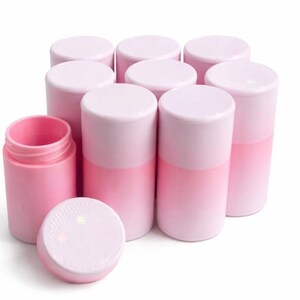 Op de afbeelding: Een verzameling roze en witte cilindervormige containers, sommige met deksels, op een witte achtergrond. Eén container is open, waardoor de binnenkant zichtbaar is. De deksels hebben een sterrenpatroon.