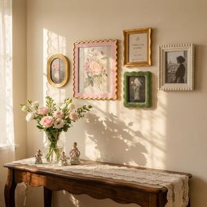 May include: A collection of four wall-mounted picture frames in oval, scalloped, and beaded styles, featuring floral art and a portrait. A glass vase with pink and white roses sits on a wooden table with a lace runner and three small figurines.