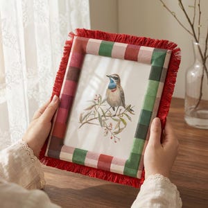 May include: A rectangular picture frame featuring a red and green checkered fabric border with a red fringe trim. The frame contains a print of a small bird with a blue throat perched on a branch with green leaves and small red berries.